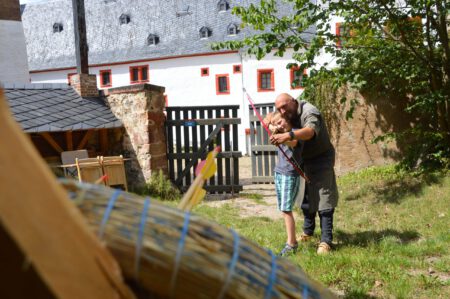 Im Bild sieht man einen als Ritter gekleideten Mann, der einem Jungen das Bogenschießen beibringt.